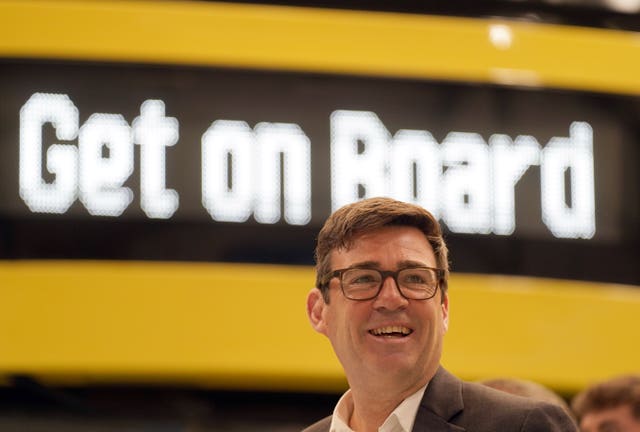 Andy Burnham smiling in front of a yellow bus with a sign reading 'Get on Board'