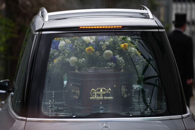 The rear of a hearse, with a coffin and funeral flowers visible through the back window