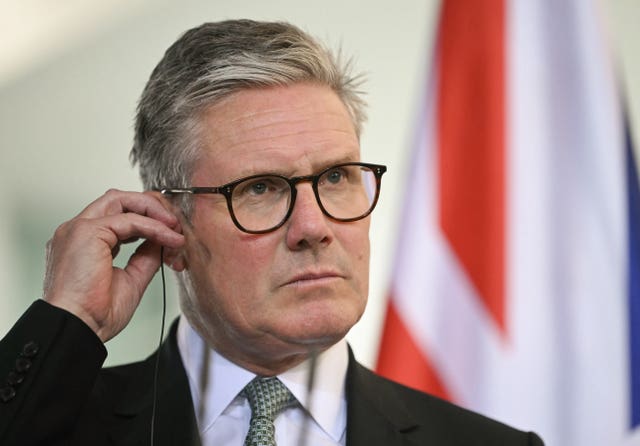 Sir Keir Starmer adjusts a headset in his left ear, a blurred Union flag can be seen in the background