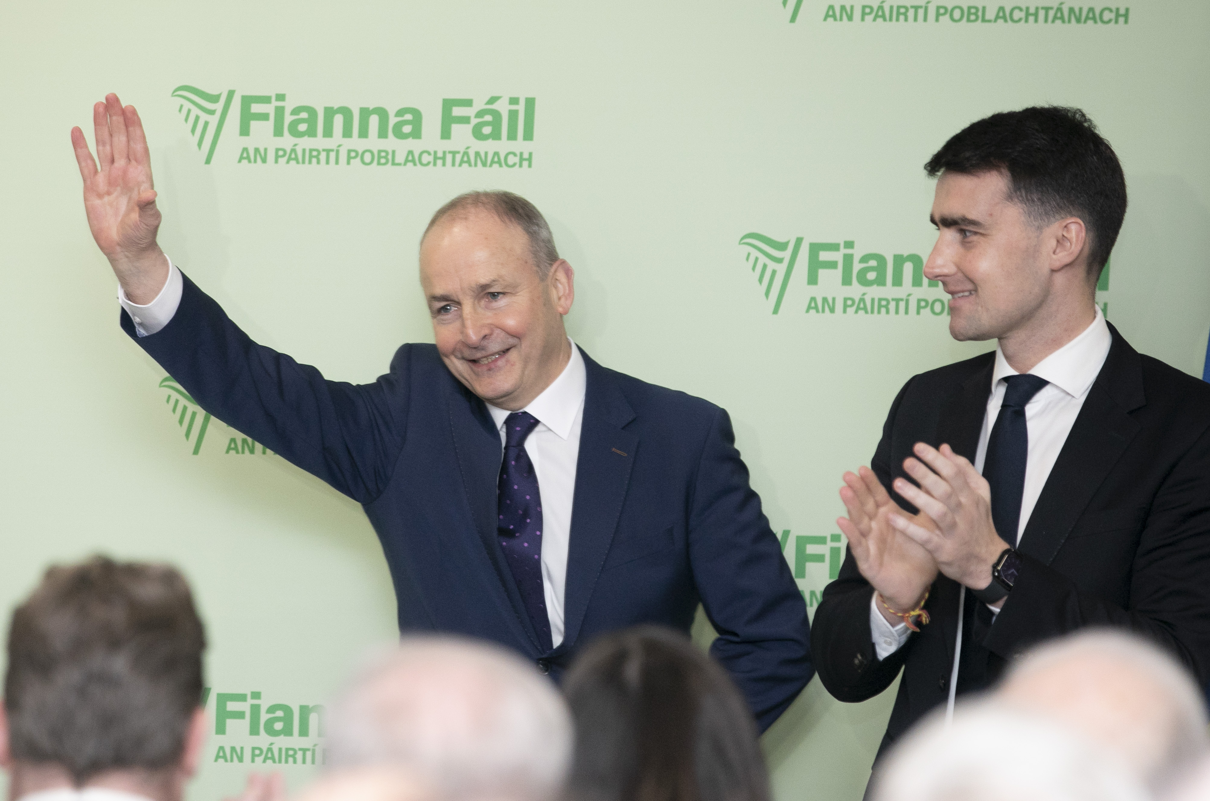 Micheal Martin smiling with one arm raised to wave, in front of Fianna Fail signage