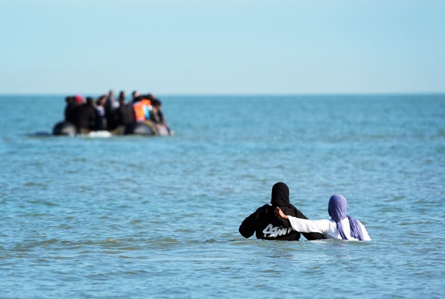 Two women in hijabs wade into chest-high water as an inflatable boat filled with migrants sails off from France