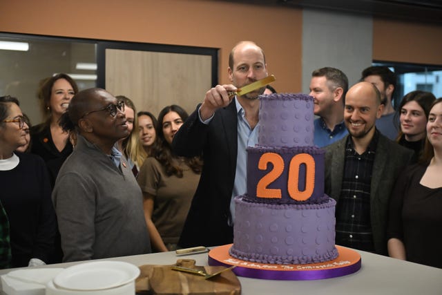 William, holding a knife, cuts into a large cake bearing the number 20 as people watch on