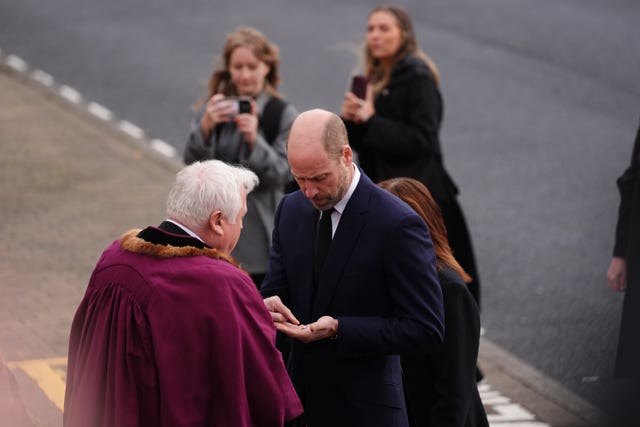 The Prince of Wales looking at a 60th anniversary pin displaying a clock face on it with the time the coal tip collapsed, which he was asked to forward on to the King, as he arrived at Aberfan and Merthyr Vale Community Centre