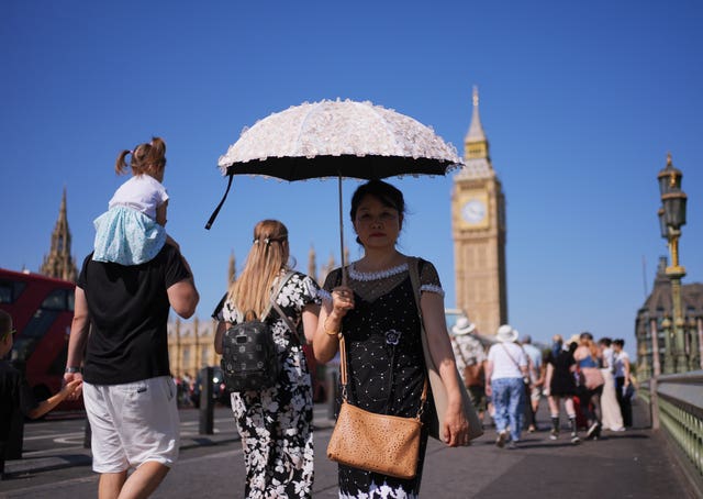 People walking along Westminster Bridge with an umbrella for shade, against the backdrop of the Houses of Parliament and Elizabeth Tower, in London.