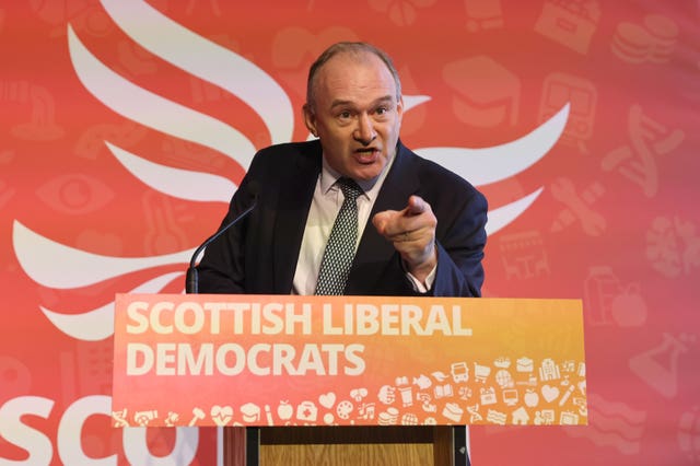 Sir Ed Davey leaning forward and pointing from behind a lectern bearing the words 'Scottish Liberal Democrats' in front of an orange backdrop featuring the Lib Dem logo