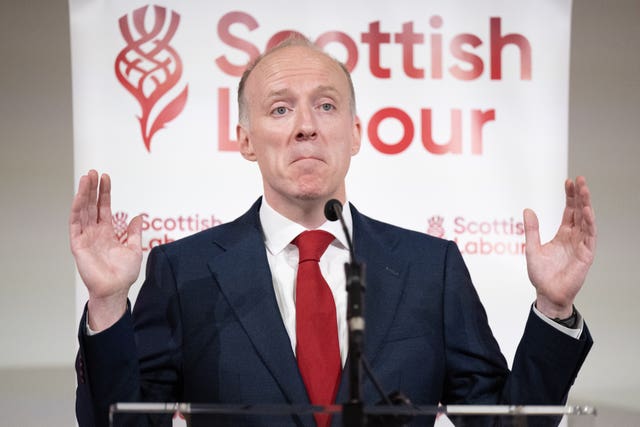 Michael Marra speaking, with both hands raised, in front of Scottish Labour signage