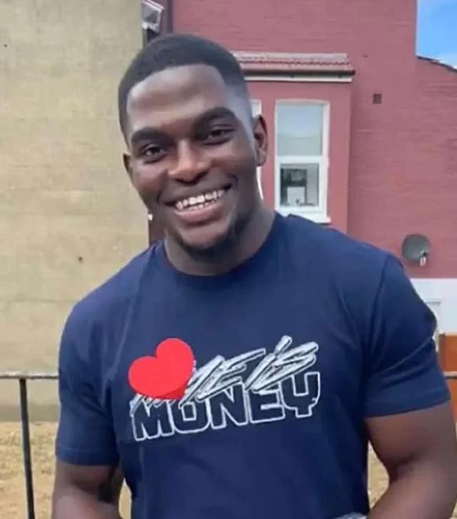 Chris Kaba smiling at the camera wearing a blue t-shirt with white writing and a red love heart on it.