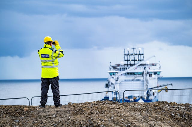 A man in hard hat and hi-vis jacket uses binoculars to look out to sea from land
