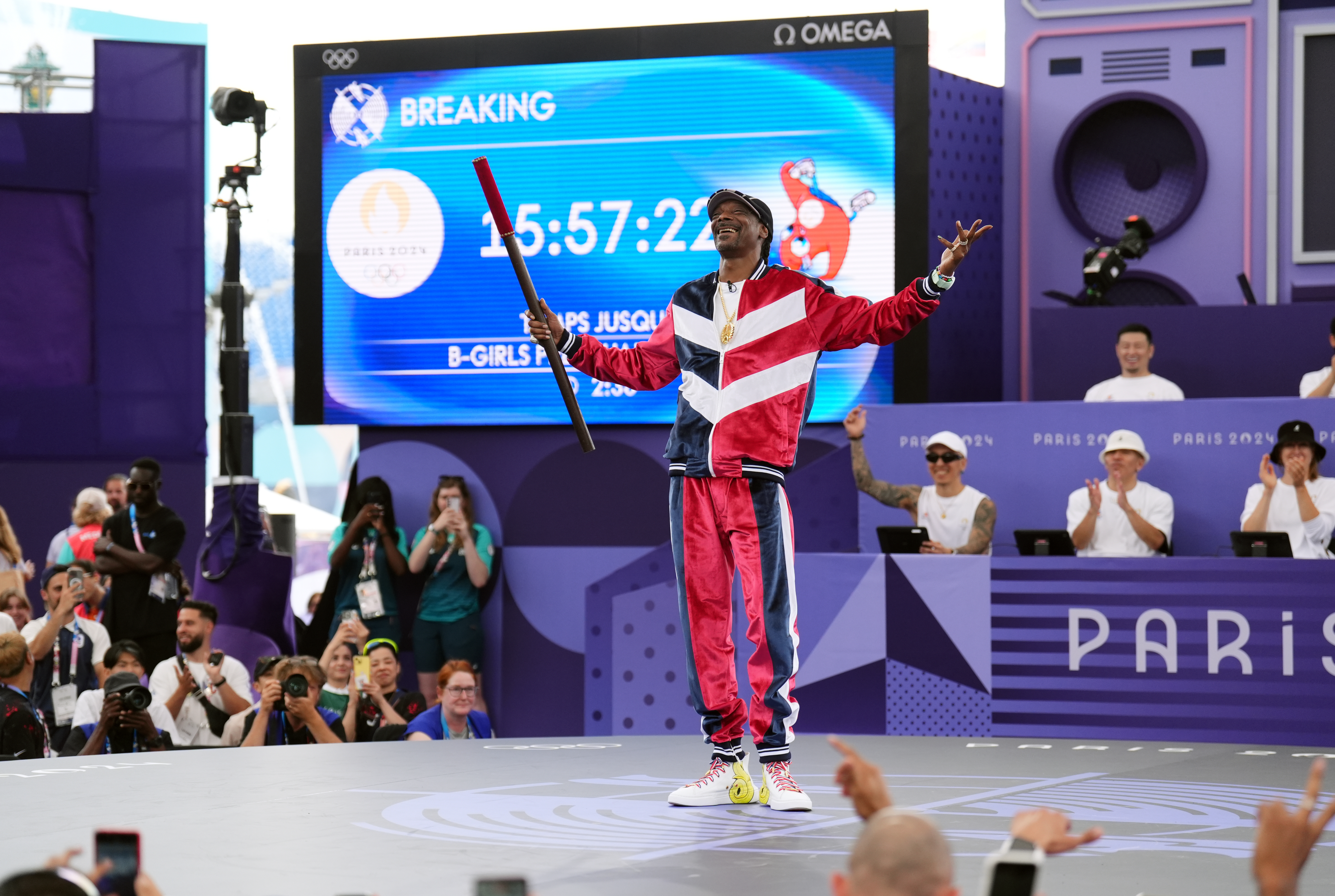 Snoop Dogg dressed in tracksuit holds his arms in the air on a stage at the Paris Olympics