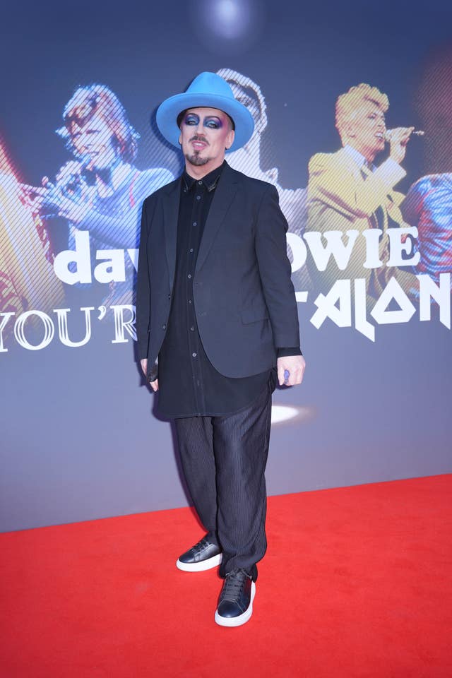 Boy George posing for photos at a red carpet, wearing a blue hat