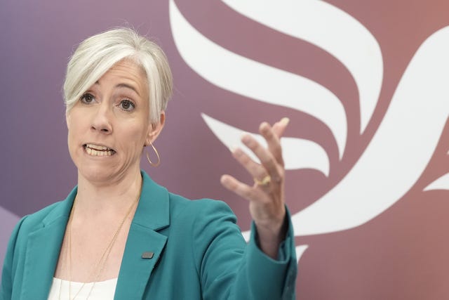 Daisy Cooper speaking, with one hand raised, in front of a large Lib Dem logo