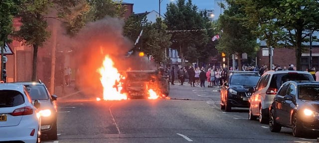 A vehicle on fire on a road, with a group of people in the background