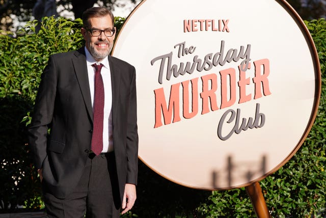 Richard Osman wearing a suit standing next to a backdrop advertising the Netflix film adaptation of The Thursday Murder Club