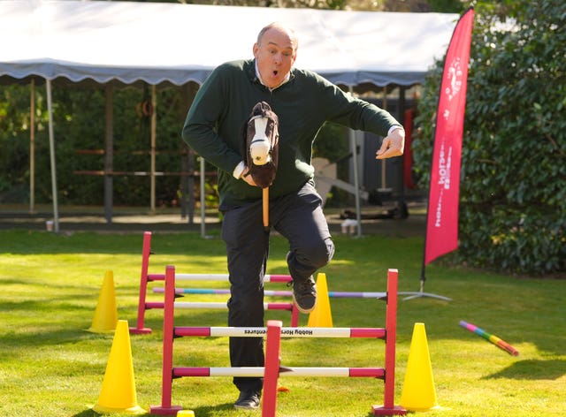 Sir Ed Davey, holding a hobby horse, jumping over a hurdle