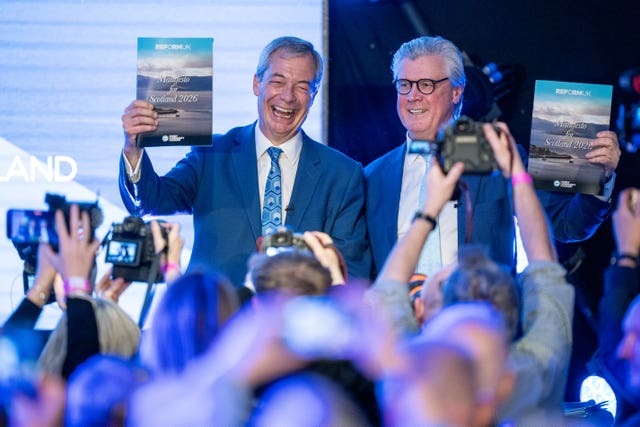 Nigel Farage and Malcolm Offord smiling while holding up copies of the Reform manifesto, in front of a bank of photographers