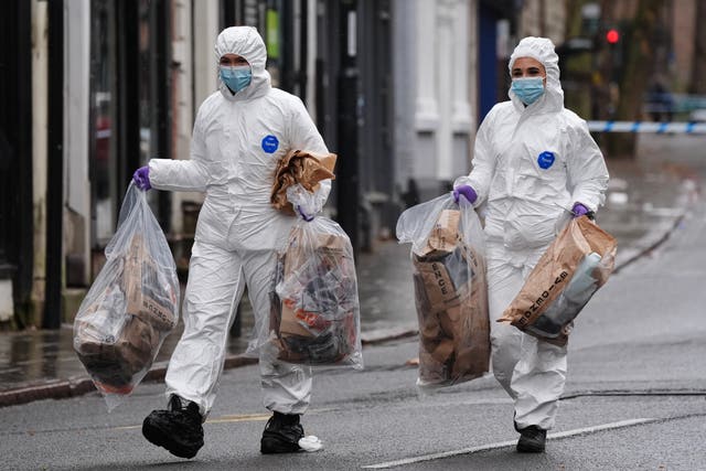 Two forensic investigators in white suits carry bags full of items along a street