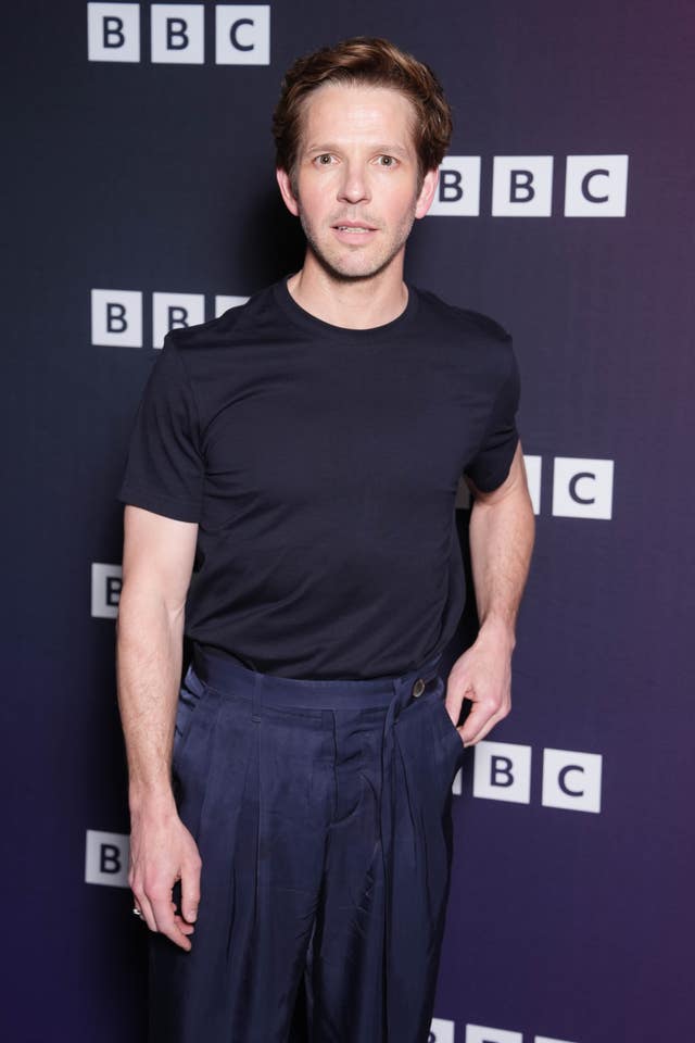 Damien Molony wearing a navy T-shirt and trousers posing in front of a blue BBC background