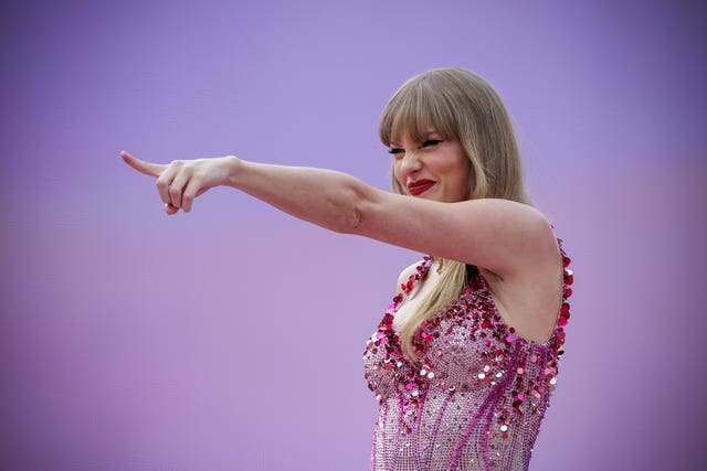 Taylor Swift smiling and pointing, wearing a pink sparkly outfit on stage