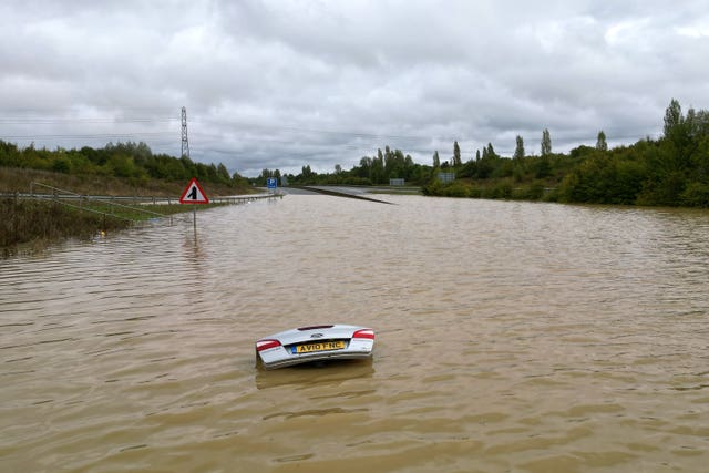 The open boot of a car is visible above the water where the vehicle is submerged in flood water