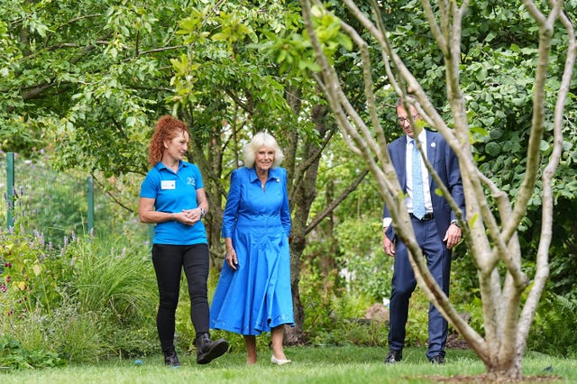 Queen Camilla in a blue dress walks through the greenery of the garden with Battersea staff on either side