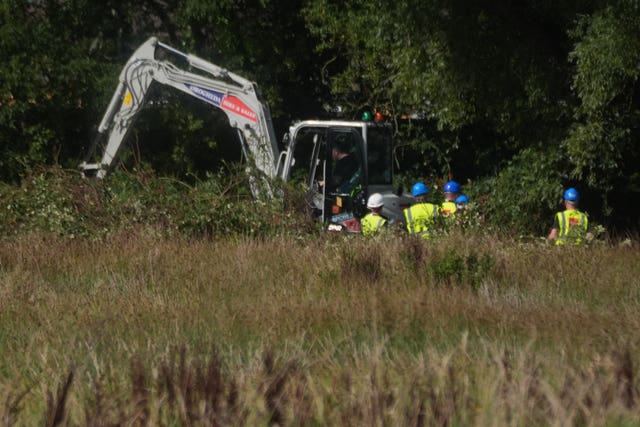 An excavator being used in long grass, with people in hard hats nearby