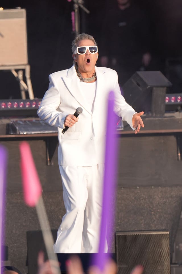 Robbie Williams dressed in a white suit and sunglasses, holding a microphone and with his mouth open wide on stage