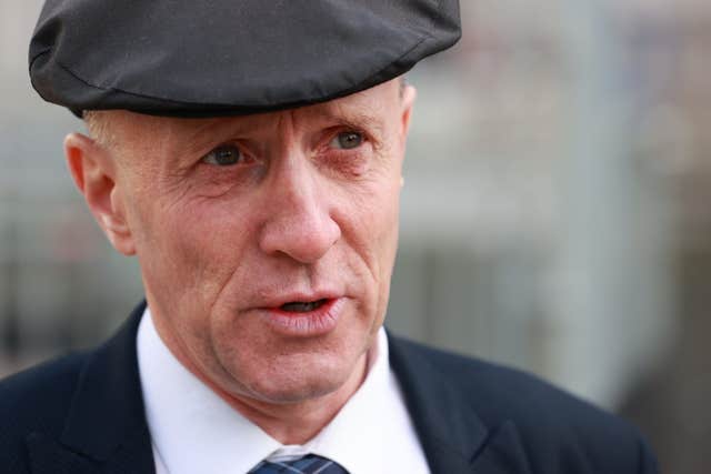 A middle-aged man wearing a dark flat cap, suit jacket, shirt, and tie looks slightly to the side in an outdoor setting