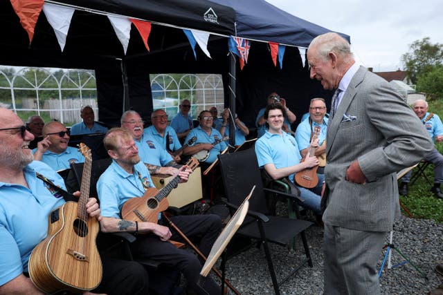 The King standing while speaking to members of a band, who are all seated and holding ukuleles