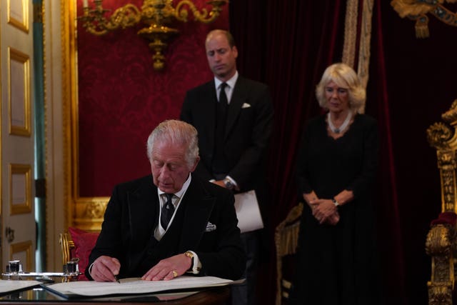 The King sits at a table and signs a document with Camilla and William standing behind him during his Accession Council at St James’s Palace in 2022