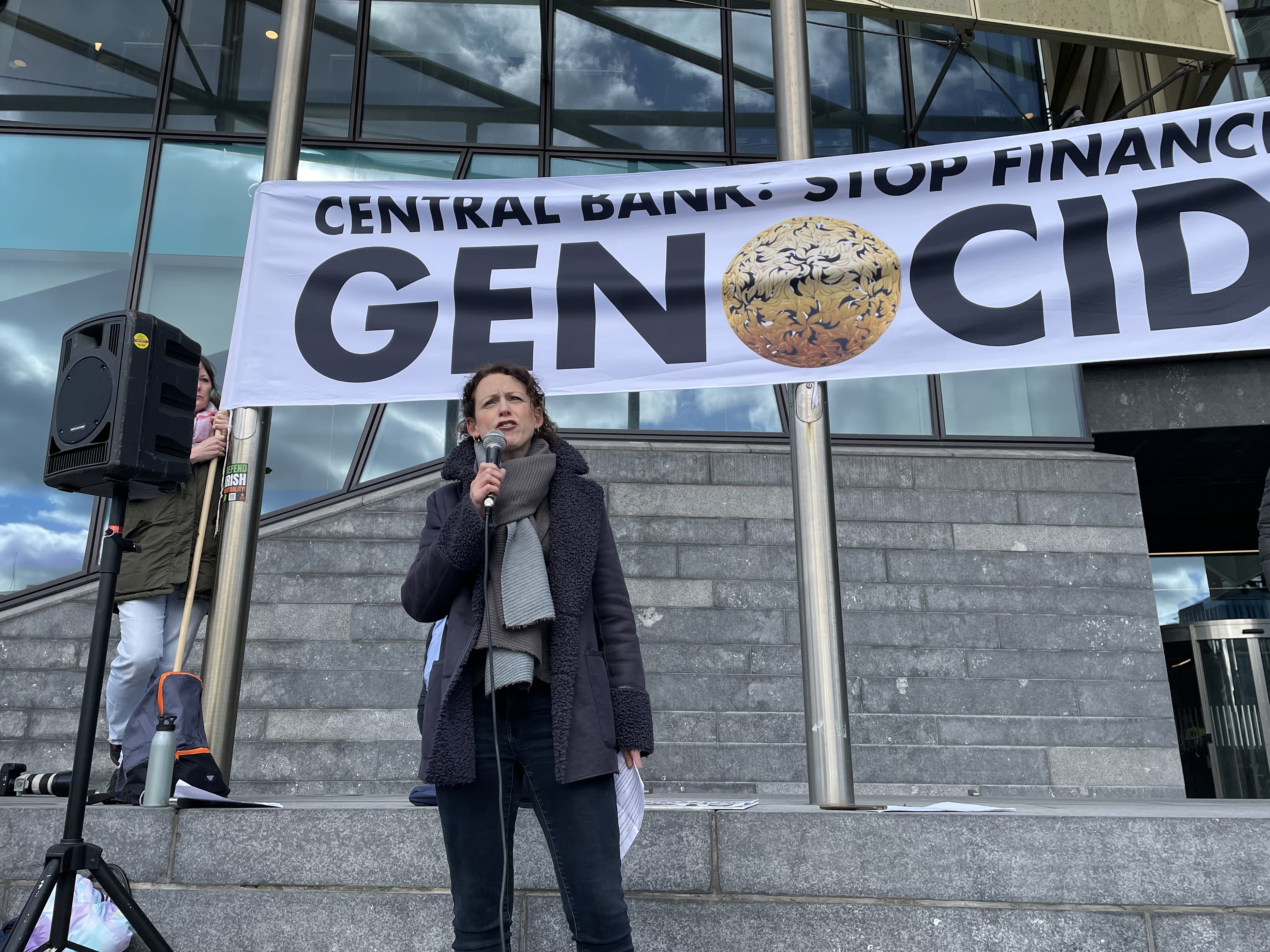 Marie Sherlock speaking into a microphone, below a banner reading 'Central Bank: Stop financing genocide'