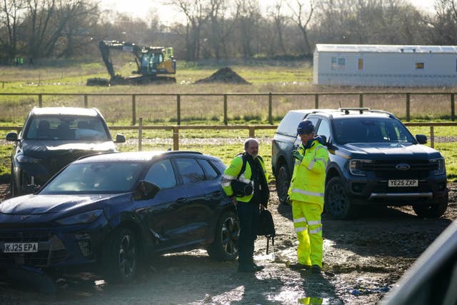 People in hi-vis gear standing next to parked cars with a digger and temporary cabin in the field beyond