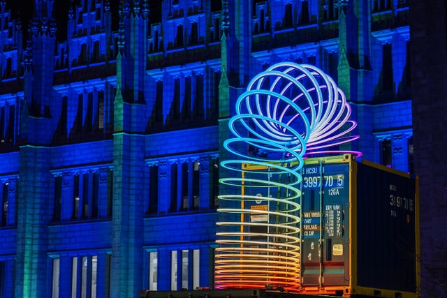 A giant illuminated slinky, on top of a contained in front of a building lit up in blue