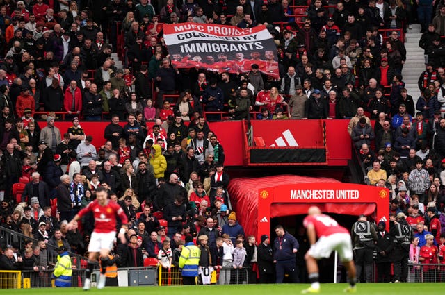 Manchester United fans in the stands hold up a banner reading MUFC Proudly Colonised By Immigrants