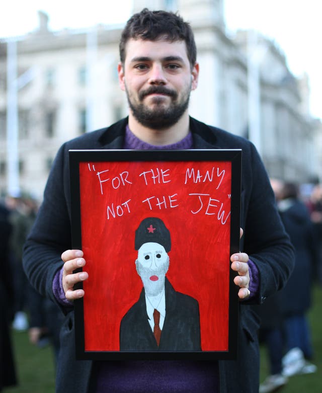 A man holds a painting of Jeremy Corbyn that says 'For the many, not the Jew'