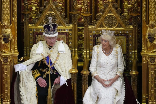 Charles and Camilla on thrones in the House of Lords with the King wearing ermine and his crown and Camilla in a white gown sitting beside him