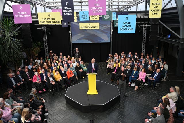 John Swinney speaking from a small platform in the middle of a room, with an audience on four sides