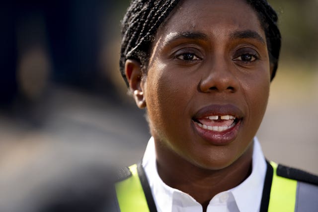 Headshot of Kemi Badenoch speaking, wearing a yellow hi-viz vest and white shirt
