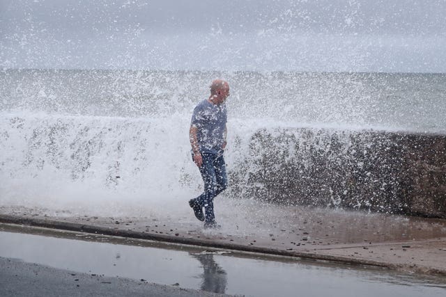 A person walking next to a sea wall, with waves splashing over it