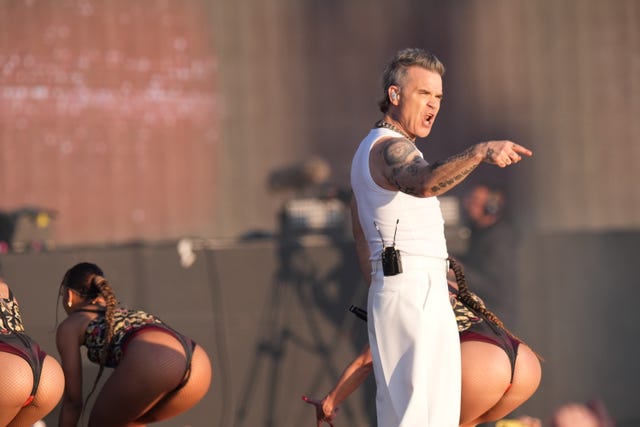Robbie Williams points out to the crowd while female dancers behind him stick out their bottoms