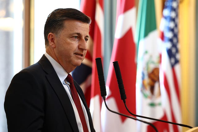 Side view of Douglas Alexander speaking into microphones, with various flags in the background