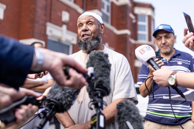 A mosque chairman stands surrounded by media with microphones and camera
