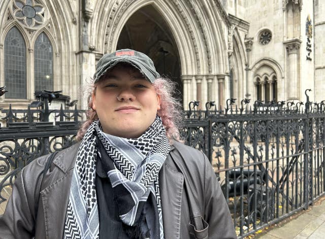 A close up of a person with pink-tinged curly hair, a leather jacket, grey baseball cap and black patterned scarf outside a stone building with black iron fencing