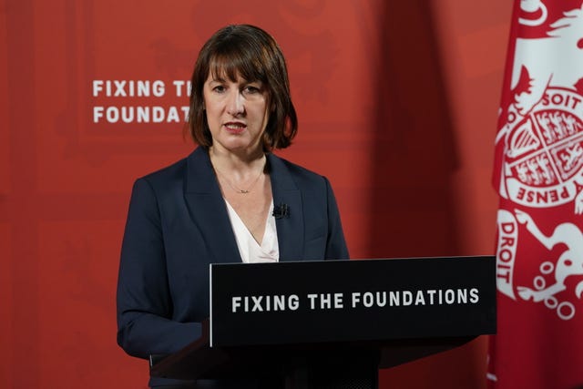 Chancellor Rachel Reeves stands behind a lectern bearing the slogan "Fixing the foundations". She stands in front of a red background and next to a red flag with a white Royal crest on it.