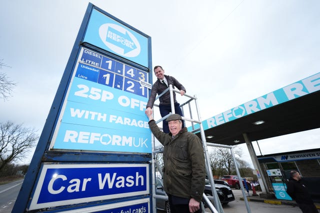 Robert Jenrick shakes hands with Nigel Farage while standing on top of scaffolding beside a petrol station pricing board covered in Reform UK signage