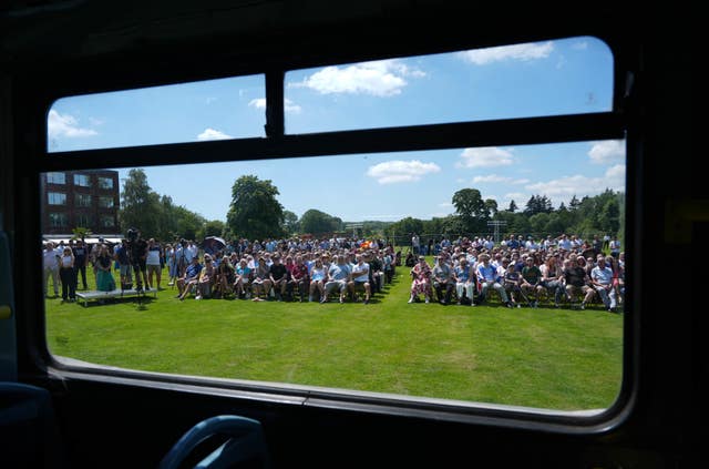 Rows of seated supporters seen through the windows of a bus with a green grass and bright blue sky background