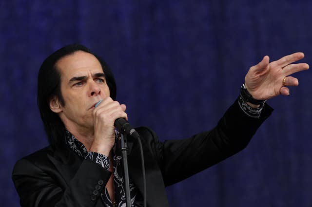 A man in a black jacket reaches out his arm while singing into a microphone