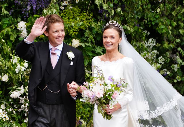 Olivia Henson and Hugh Grosvenor, wearing wedding outfits, wave to the crowd