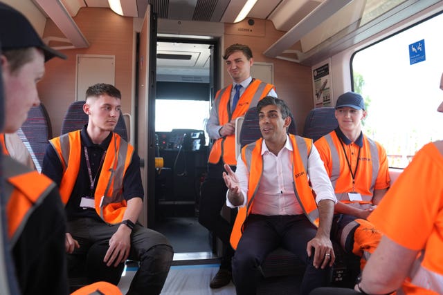 Rishi Sunak in a sleeveless hi-vis jacket seated in a train carriage talking to apprentices