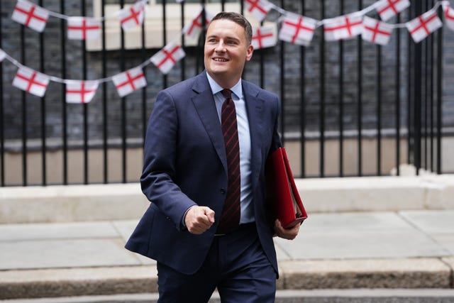 Wes Streeting walking with a red box under one arm. There's bunting of the England flag on the railings behind him