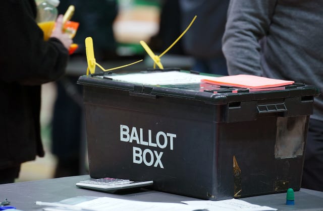 An unopened black ballot box sitting on a table, with count staff in the background